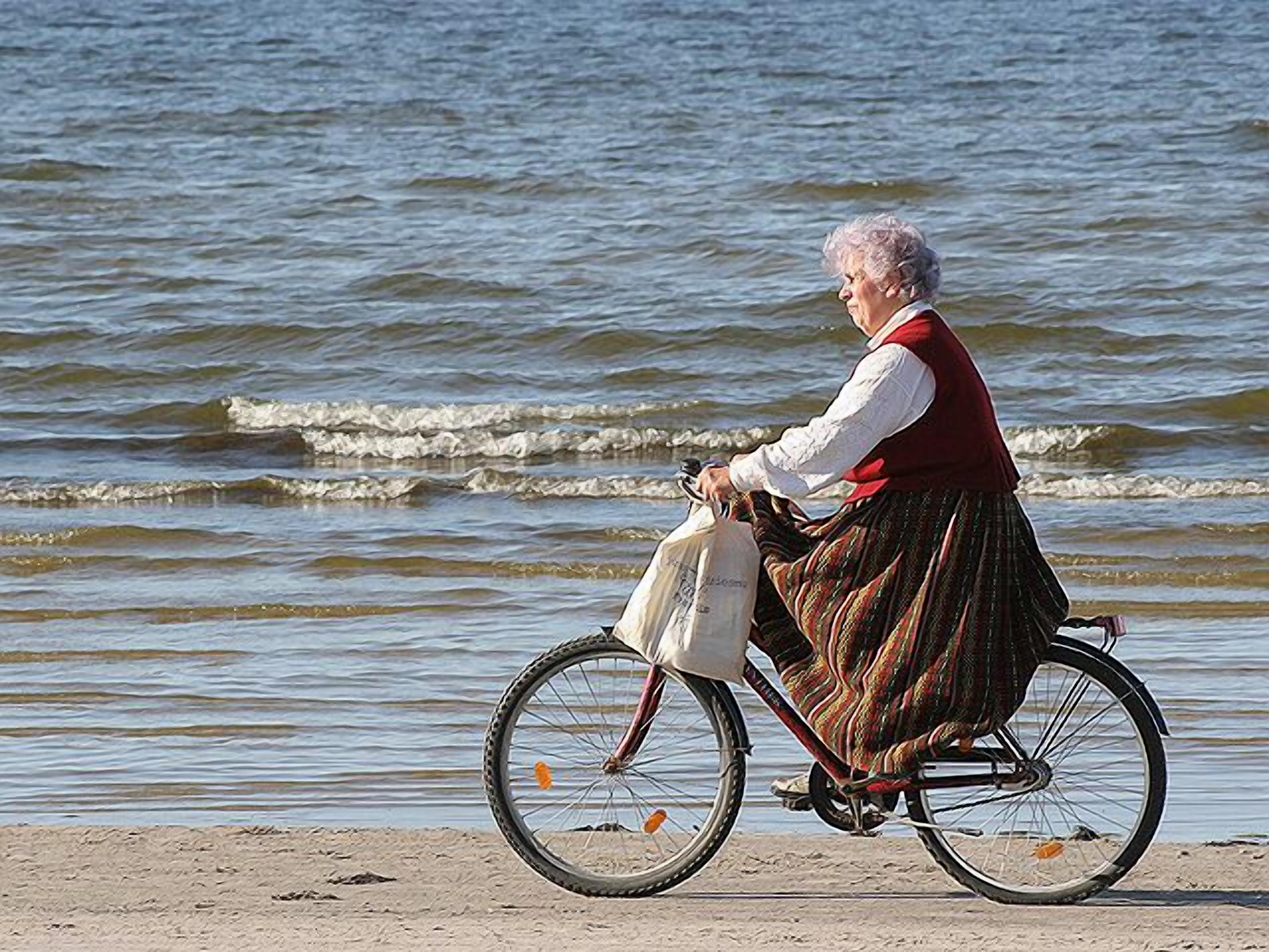 An elderly person riding a bicycle on a sandy beach with waves in the background.