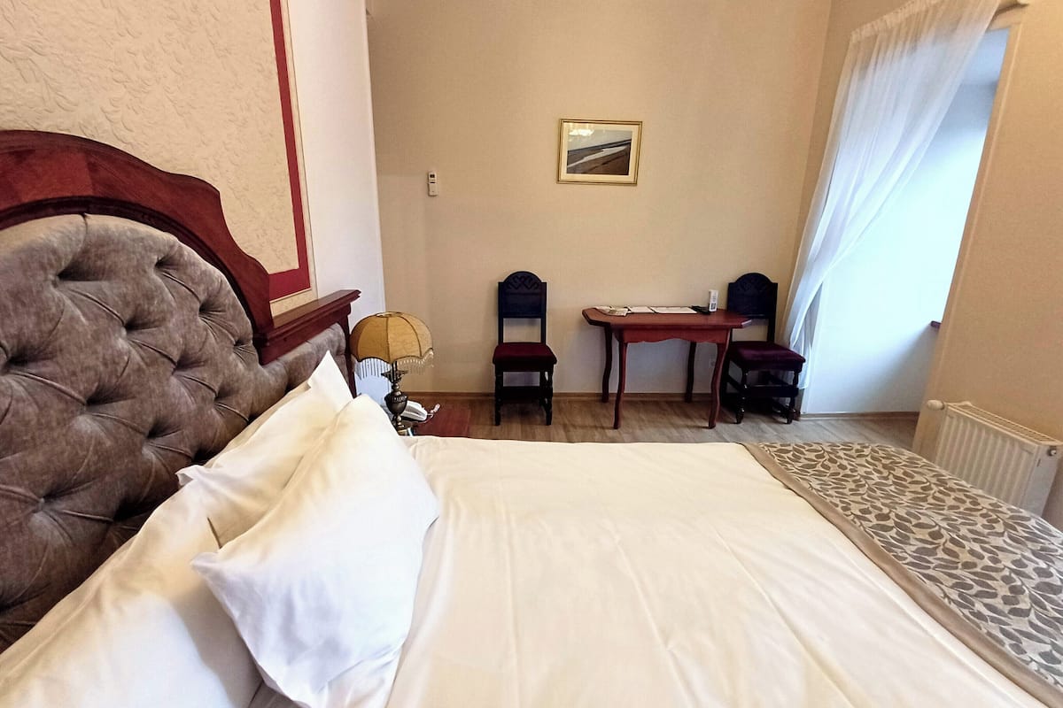 Et hotelværelse med en stor seng, et træpanel bag hovedgæret, et skrivebord med to stole, et lille bord og et billede på væggen.