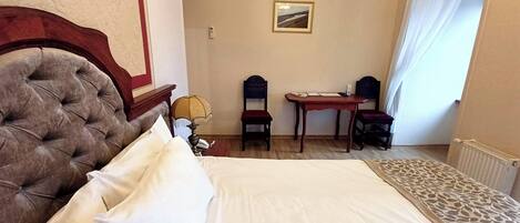 Habitación de hotel con una cama grande, cabecera de madera, escritorio con dos sillas, una mesita y un cuadro en la pared.