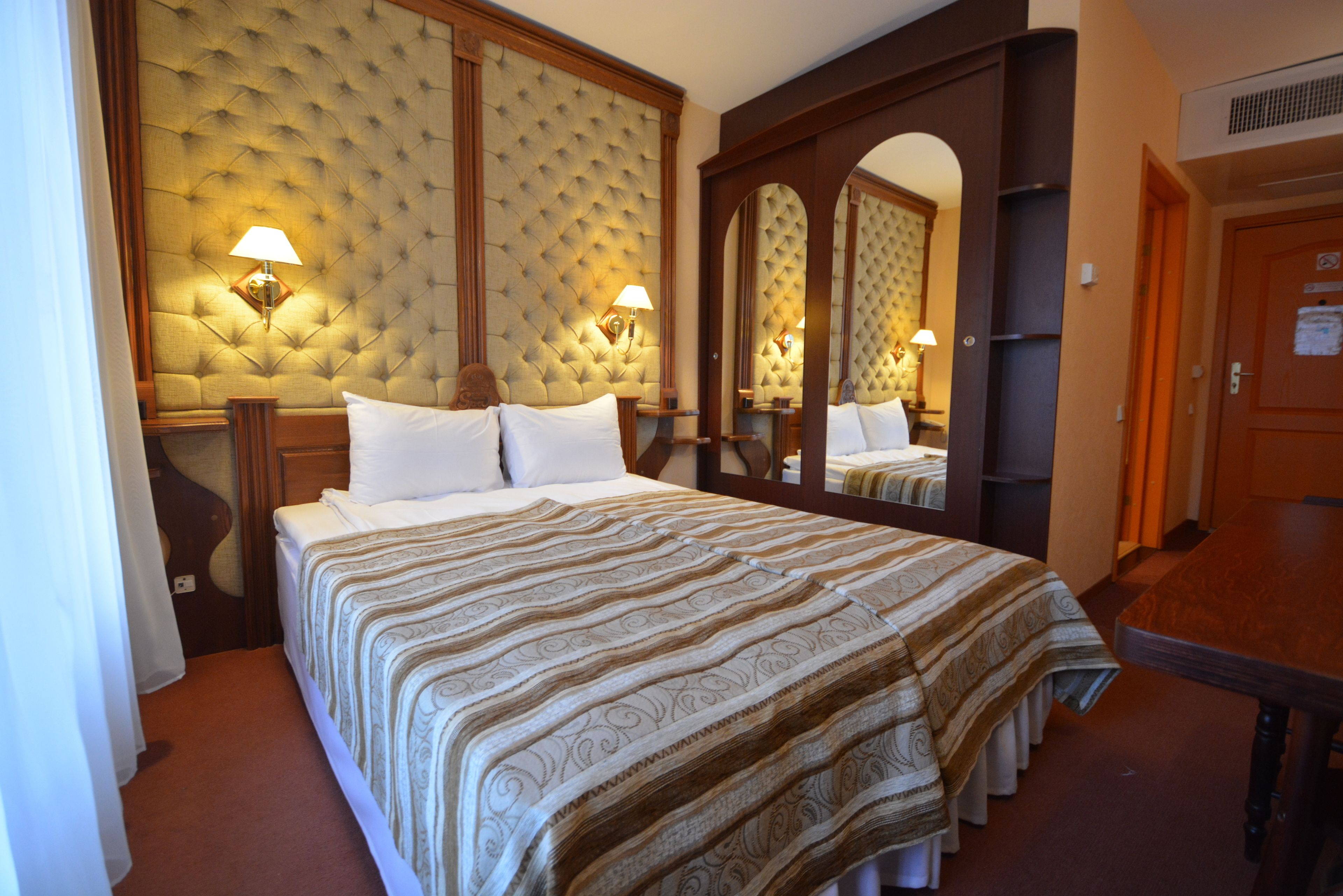 A hotel room with a large bed, a tufted headboard, two bedside lamps, a mirror, and a wooden door.