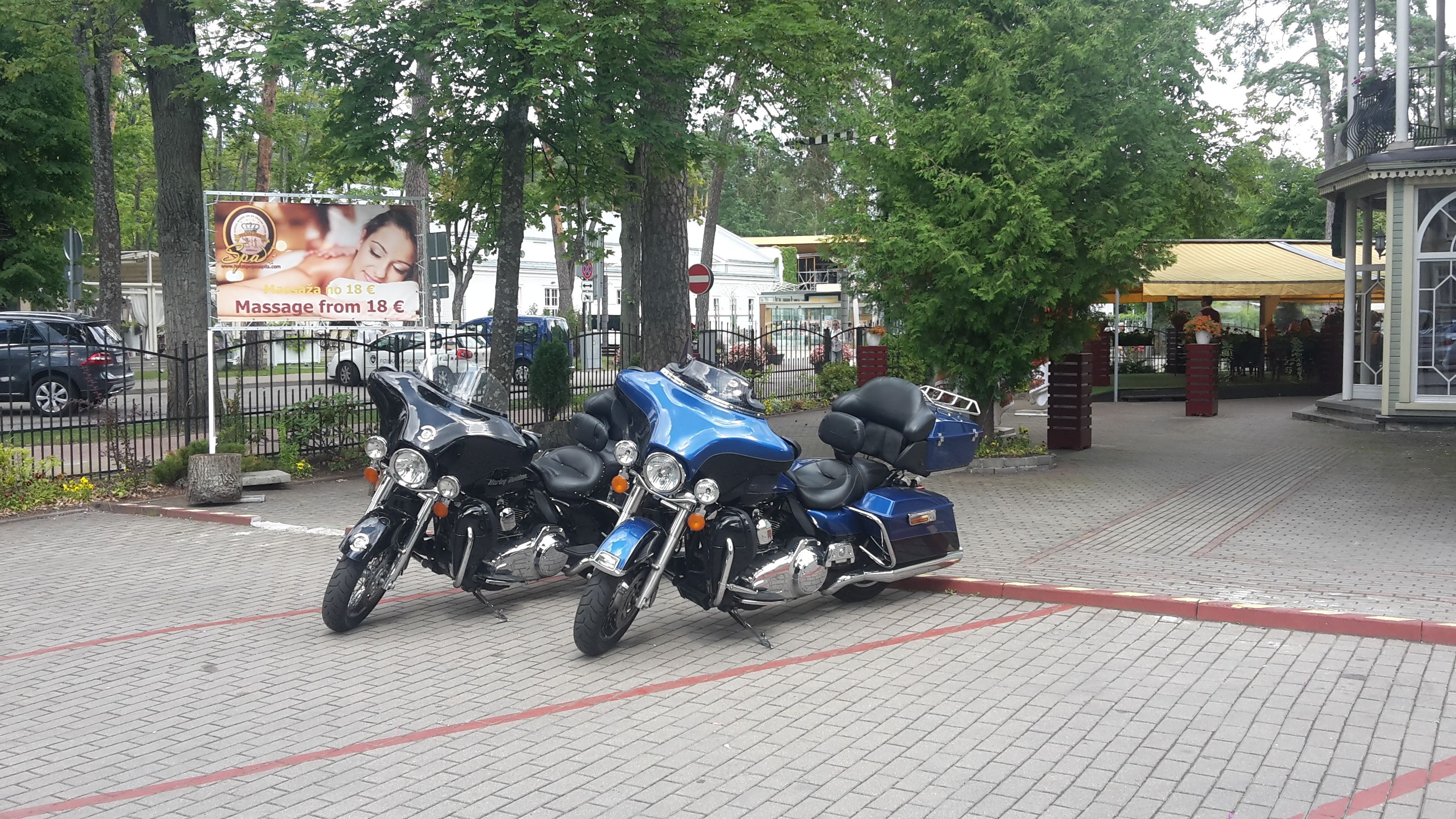 Dos motocicletas estacionadas en un área pavimentada, con árboles y un edificio al fondo.