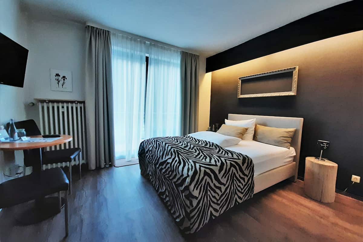Une chambre d’hôtel avec un lit, un bureau, une chaise, un radiateur et une fenêtre avec des rideaux.