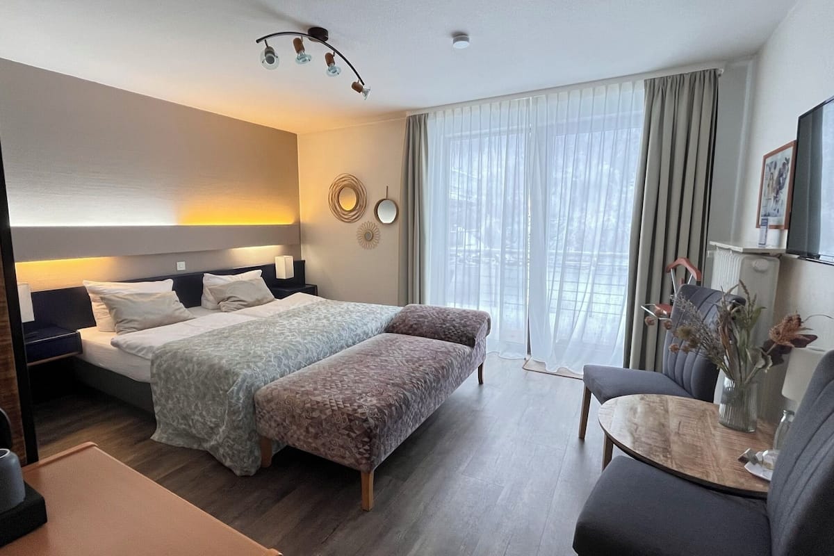 Une chambre d’hôtel avec un grand lit, un bureau avec une chaise, une petite table et une grande fenêtre avec des rideaux.