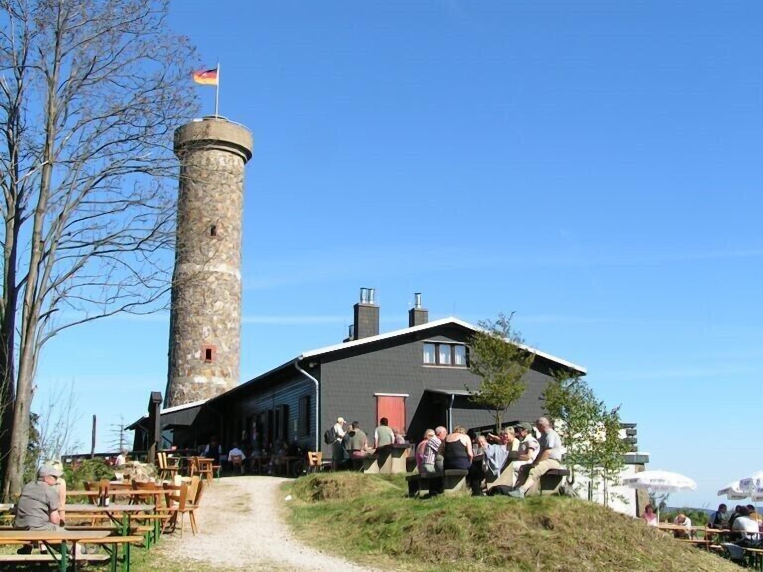 A stone tower with a flag, a building with a sloped roof, and outdoor seating with people.