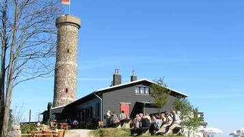A stone tower with a flag, a building with a sloped roof, and outdoor seating with people.