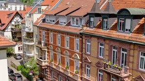 Uma fileira de edifícios europeus tradicionais com telhados de telha vermelha, uma rua com carros estacionados e uma placa indicando um hotel.