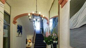 Um corredor com escada, um candelabro, uma cadeira e um vaso com flores.