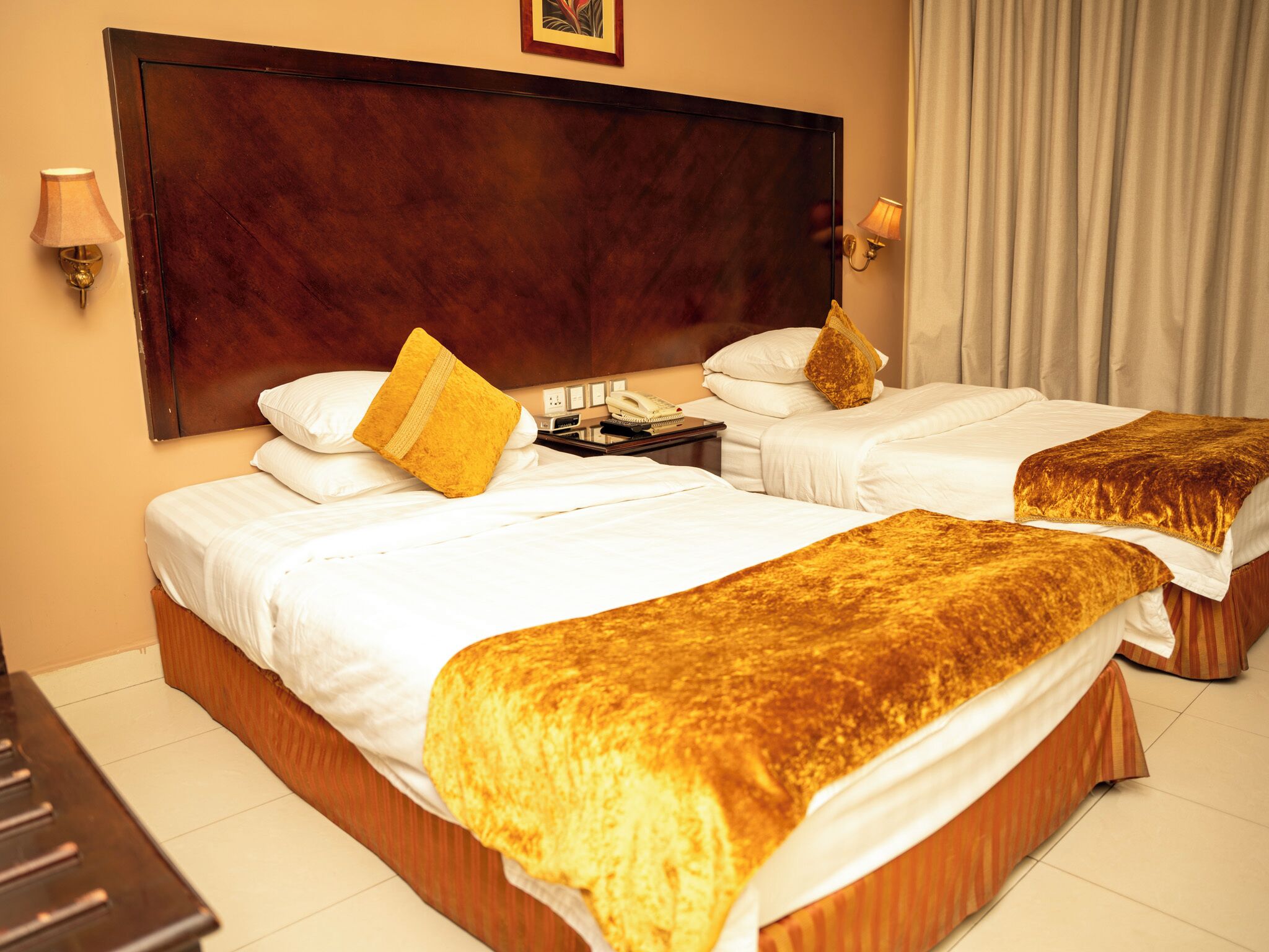 Une chambre d’hôtel avec deux lits, une grande tête de lit en bois, une table de chevet avec une lampe et une fenêtre avec des rideaux.