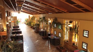 A corridor with wooden ceiling beams, potted plants, and tables and chairs arranged along the sides. - Captain's Hotel (Aqaba)