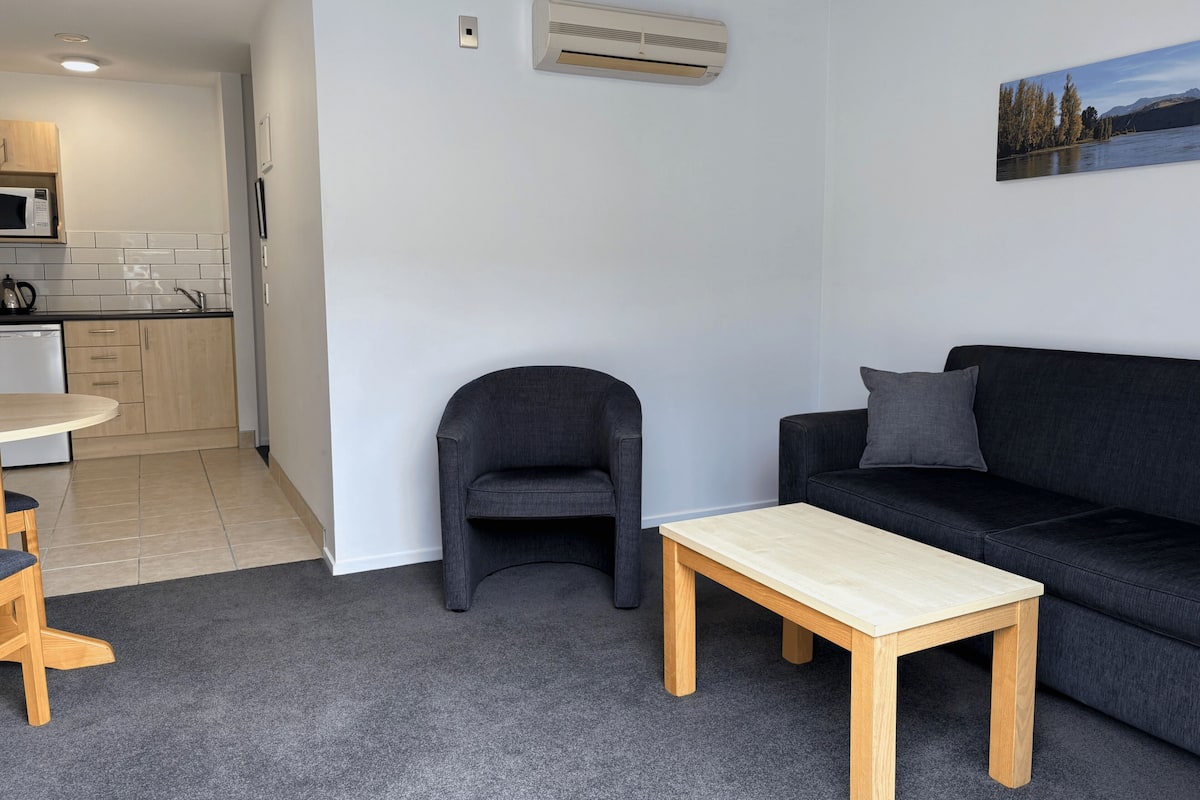 Una sala moderna con un sofá negro, una mesa de centro de madera, un sillón gris y una zona de cocina con microondas y armarios.