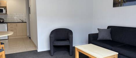 A modern living room with a black sofa, a wooden coffee table, a grey armchair, and a kitchen area with a microwave and cabinets.