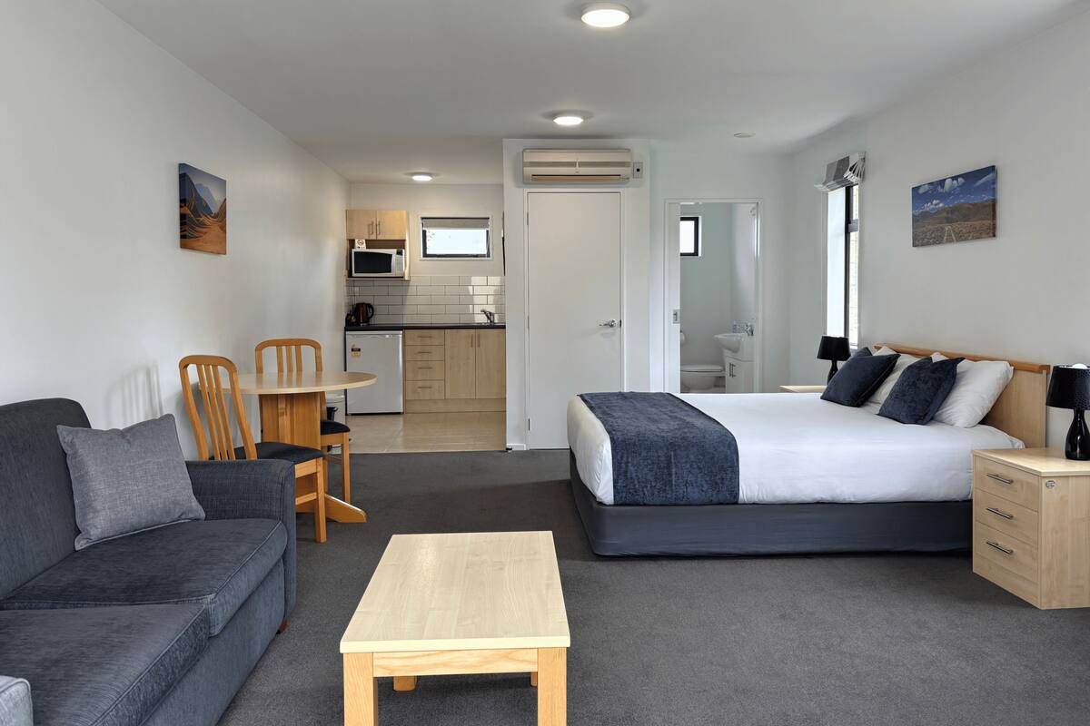 Una habitación moderna con una cama, un sofá, una mesita y una kitchenette al fondo.
