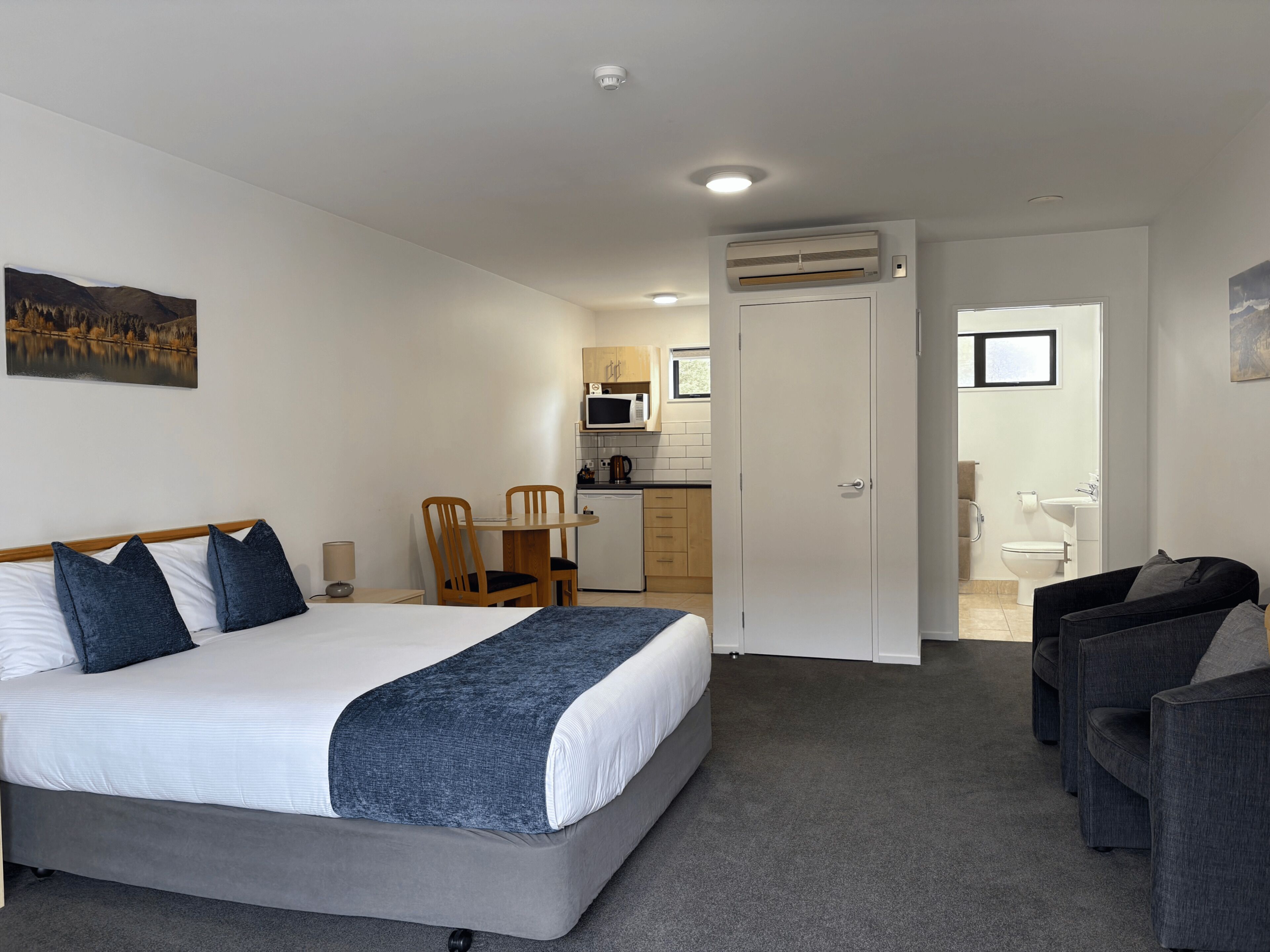 A hotel room with a bed, a desk, a chair, a kitchenette, and a bathroom.