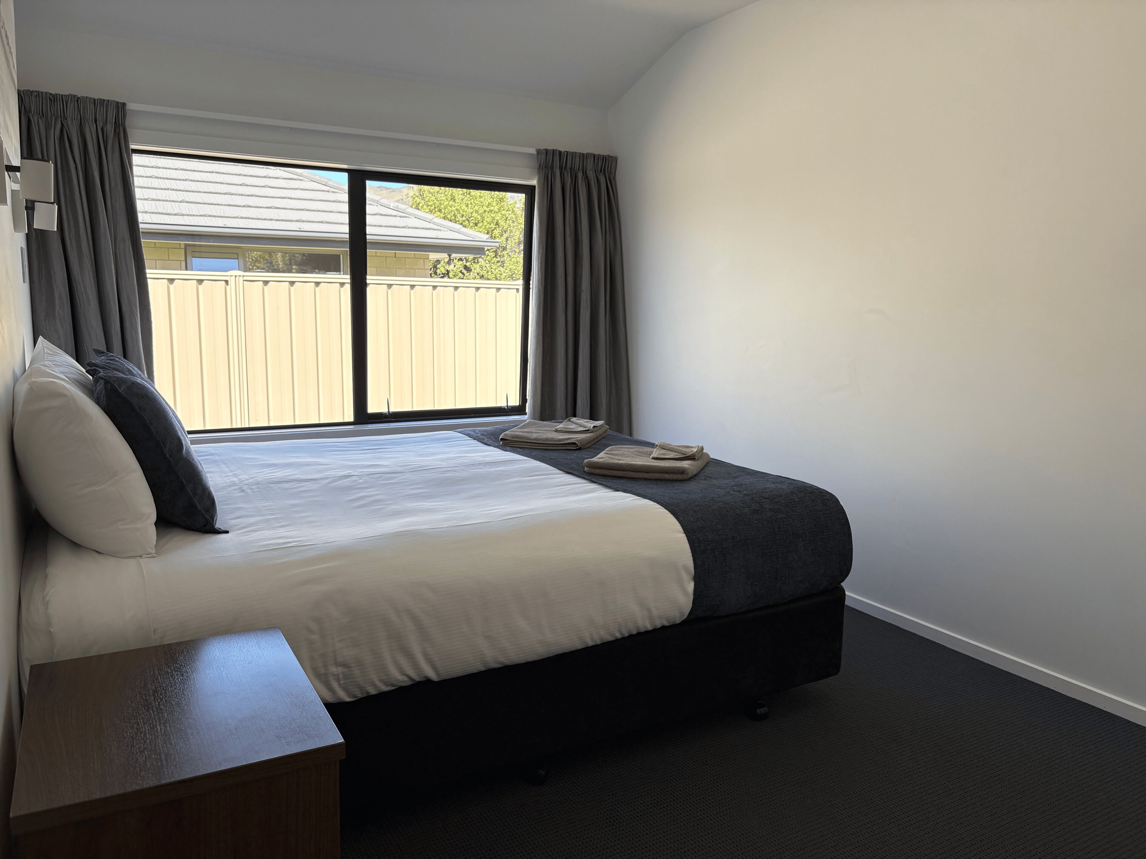 A bedroom with a bed, a bedside table, a window with curtains, and a view of a fence outside.