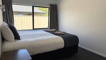 A bedroom with a bed, a bedside table, a window with curtains, and a view of a fence outside.