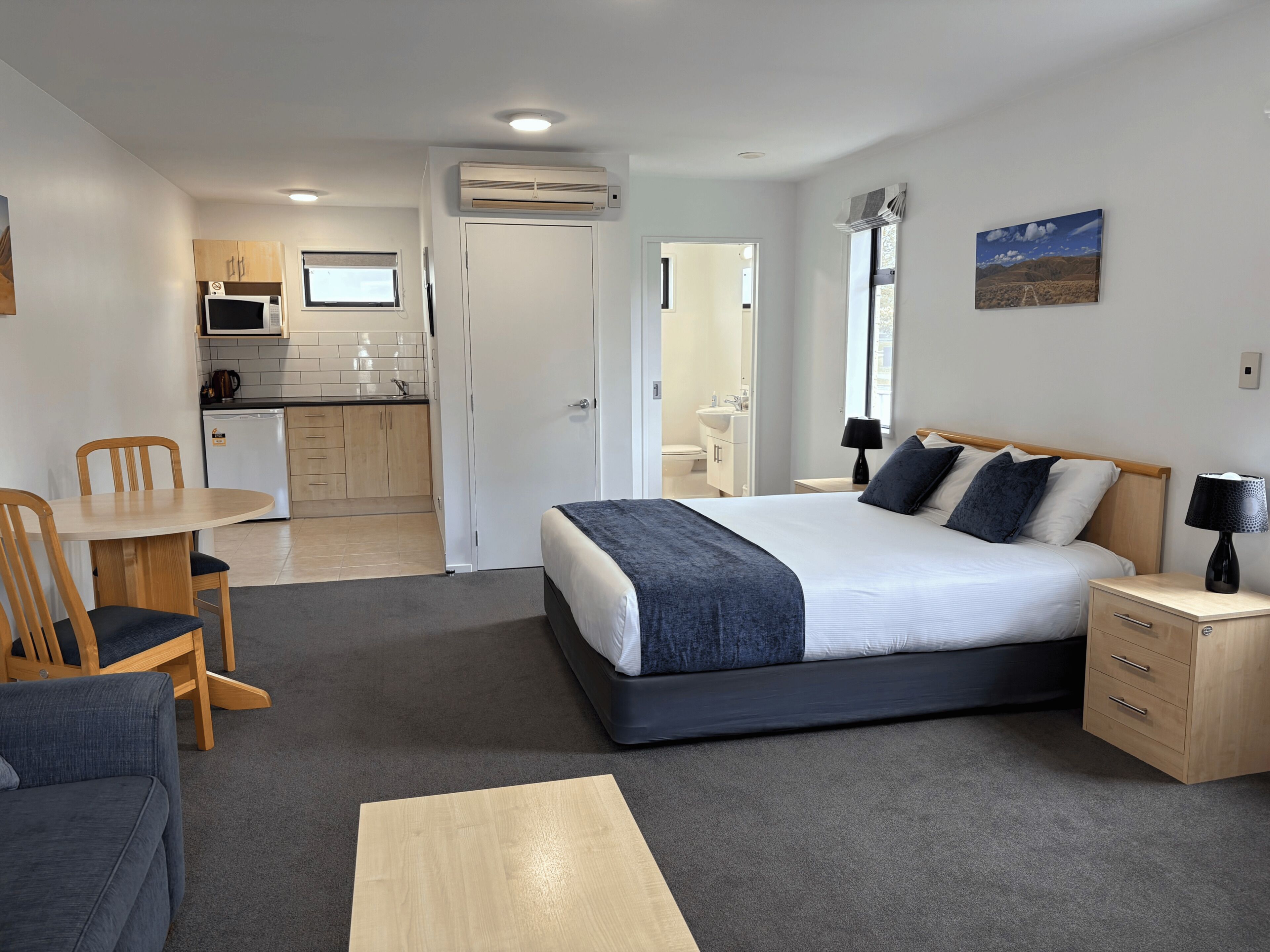 A compact hotel room with a bed, bedside table, lamp, and a view of the kitchen area.