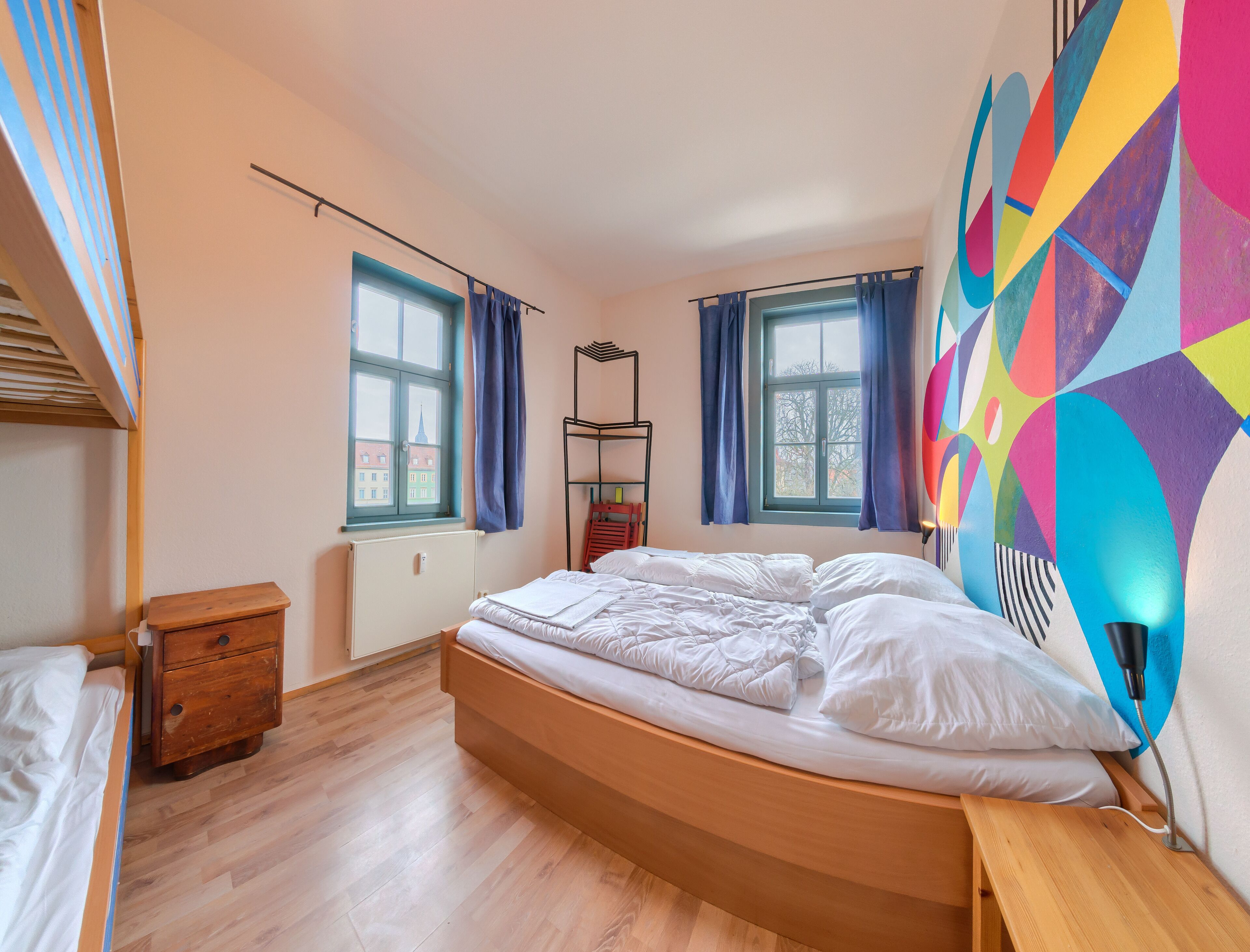 A bedroom with a wooden bed, two windows with curtains, a radiator, and a colorful wall mural.