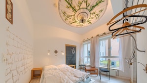 A bedroom with a bed, a desk, and a chair. There is a window with curtains, a wall with writing, and a ceiling fan. - Labyrinth Hostel (Weimar)