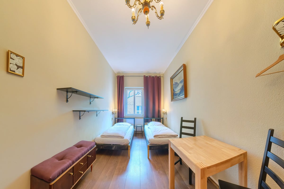 A small, well-lit room with two single beds, a wooden table, a bench, and a hanging light fixture.