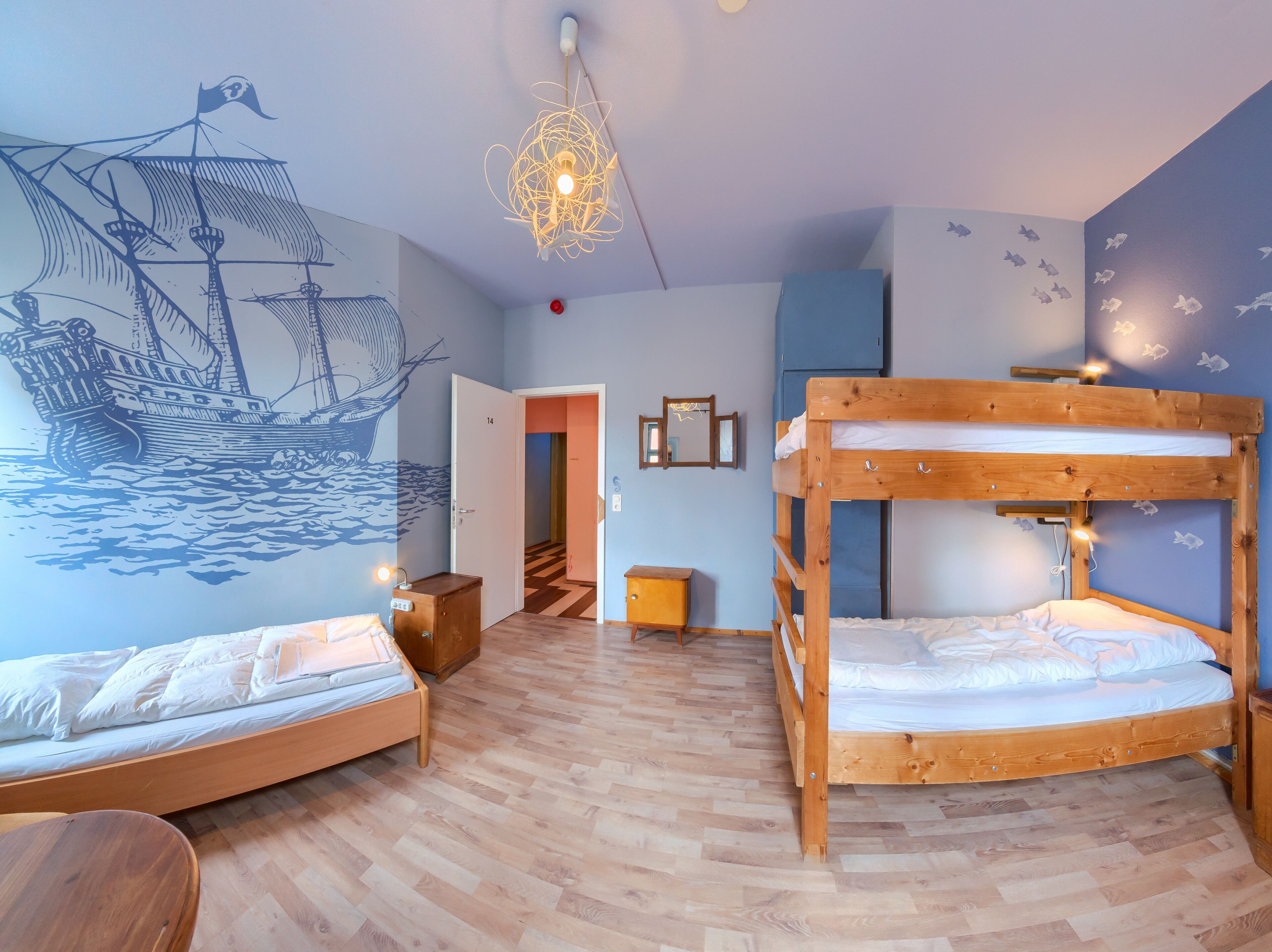 A room with two bunk beds, a wooden desk, a chair, a wall mural of a ship, and a hanging light fixture.