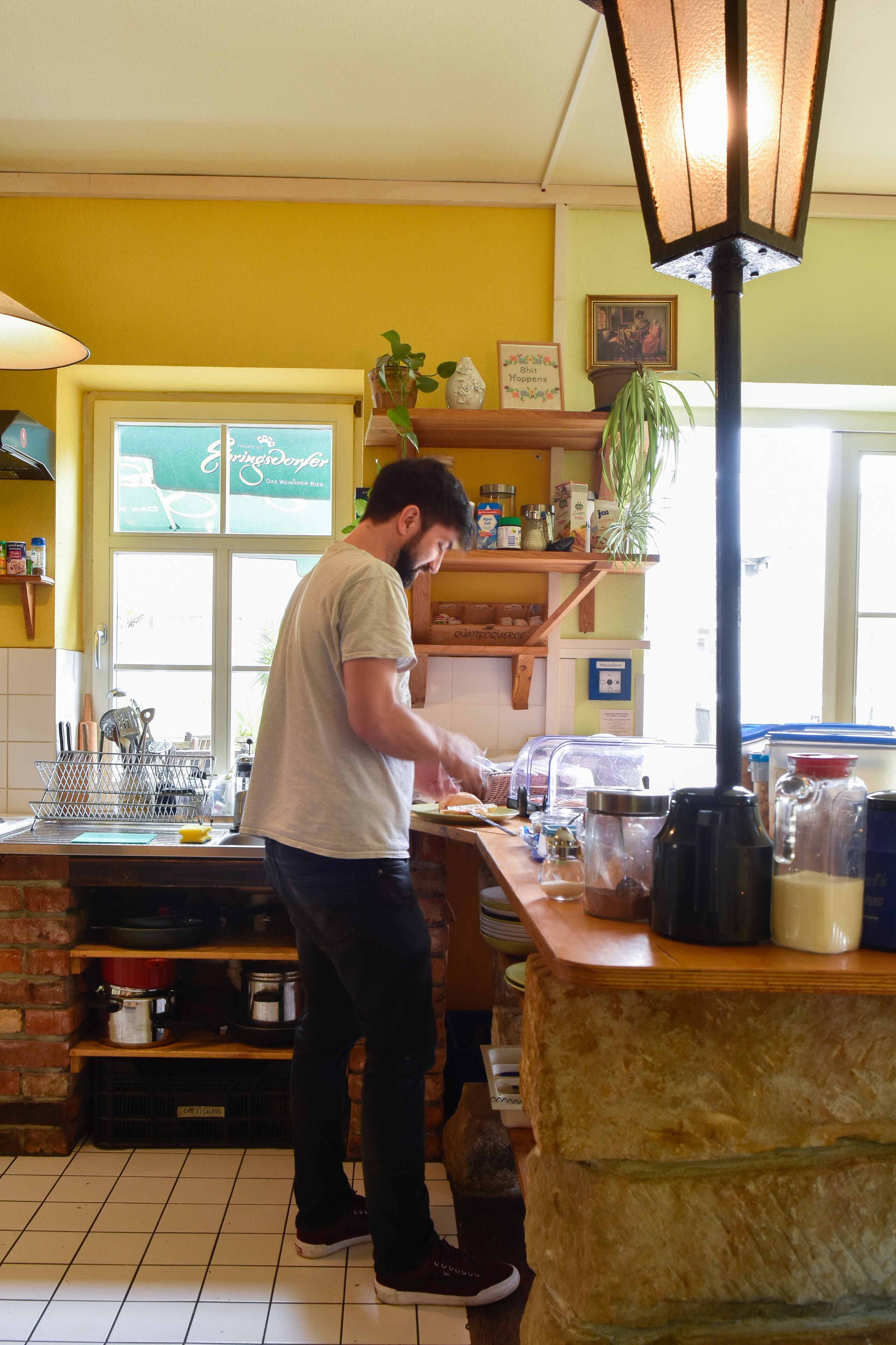 A kitchen with a person cooking, a window, shelves with various items, and a lamp.