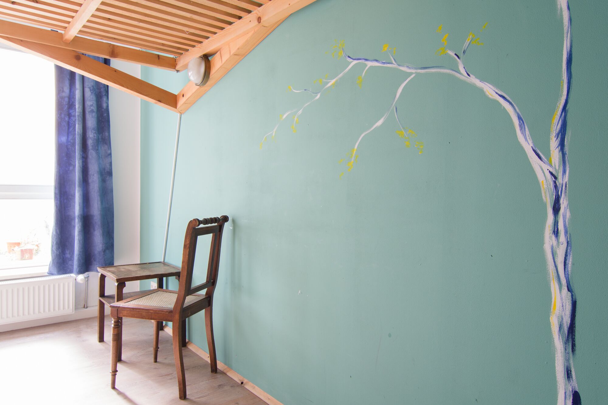A room with a wooden chair and a table, a green wall with a tree branch mural, and a window with blue curtains.