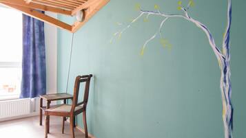 A room with a wooden chair and a table, a green wall with a tree branch mural, and a window with blue curtains.