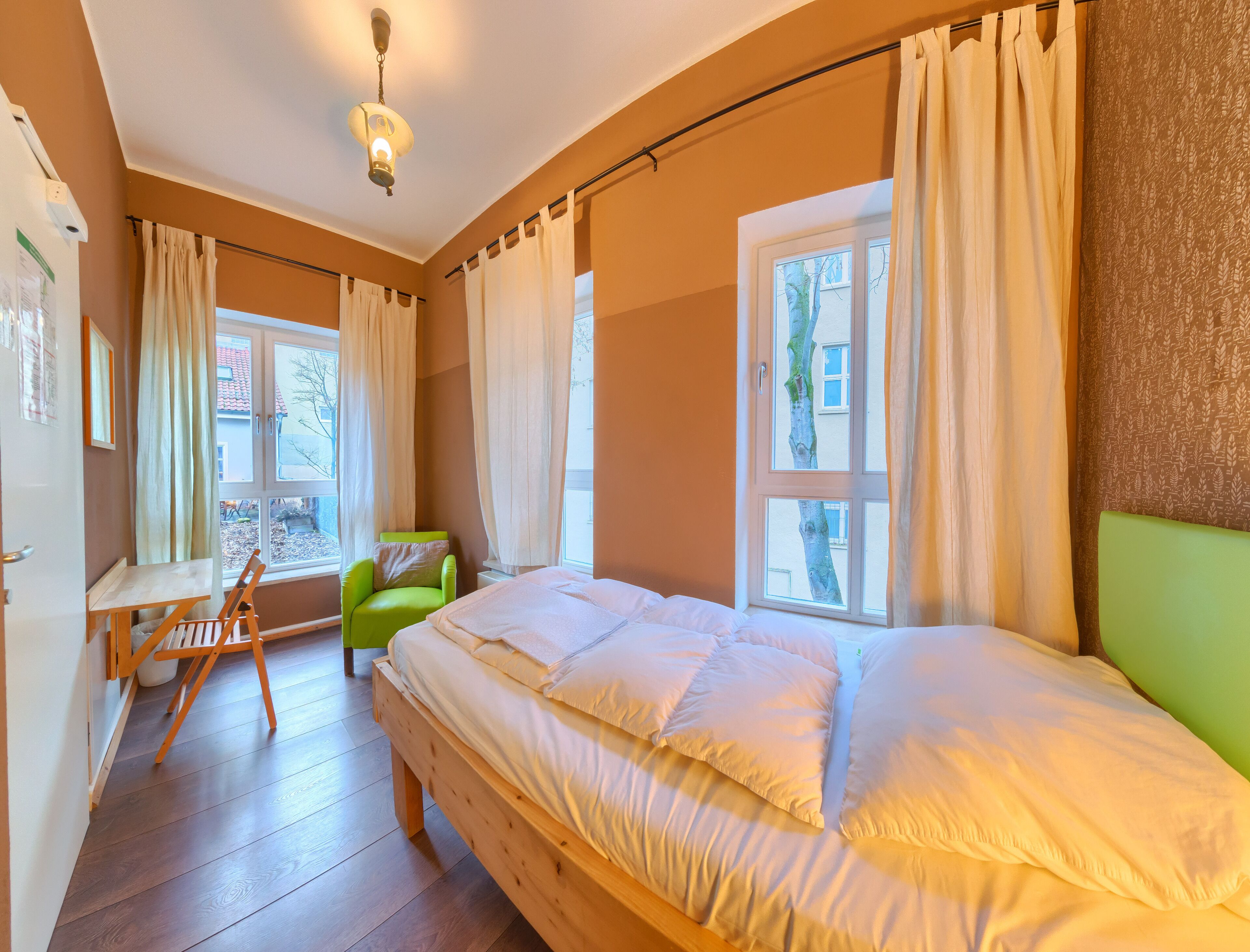 Quarto com cama de madeira, uma cadeira verde, janela com cortinas e luminária no teto.