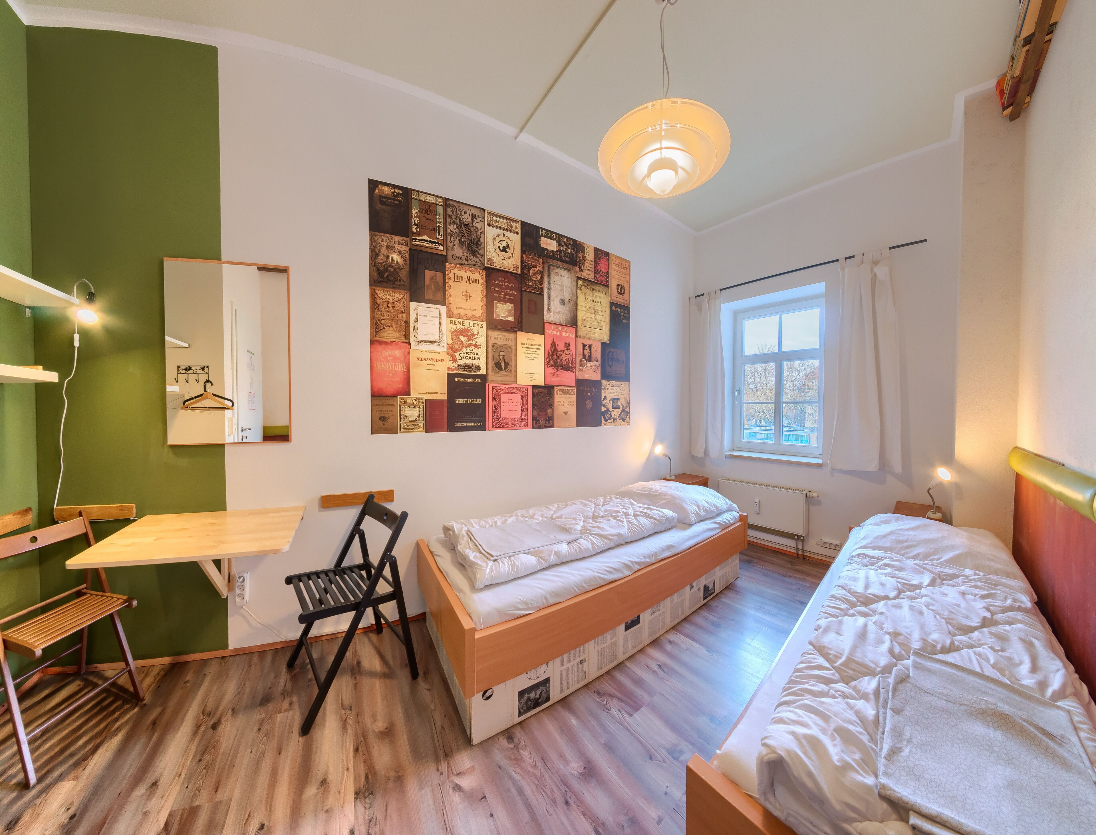 A room with a bunk bed, a desk, a chair, a wall with a collage of vintage book covers, a hanging light fixture, and a window with curtains.
