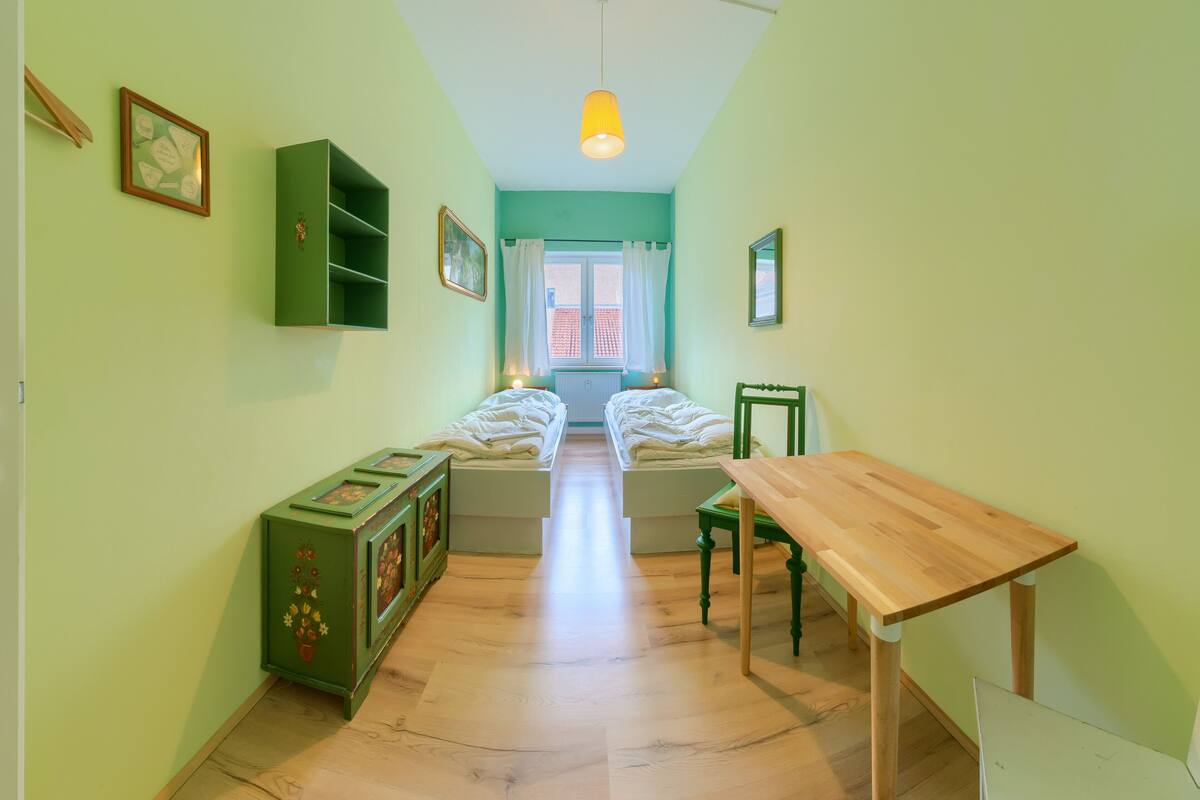 A room with two beds, a wooden table, a green cabinet, and a window with a view of buildings outside.
