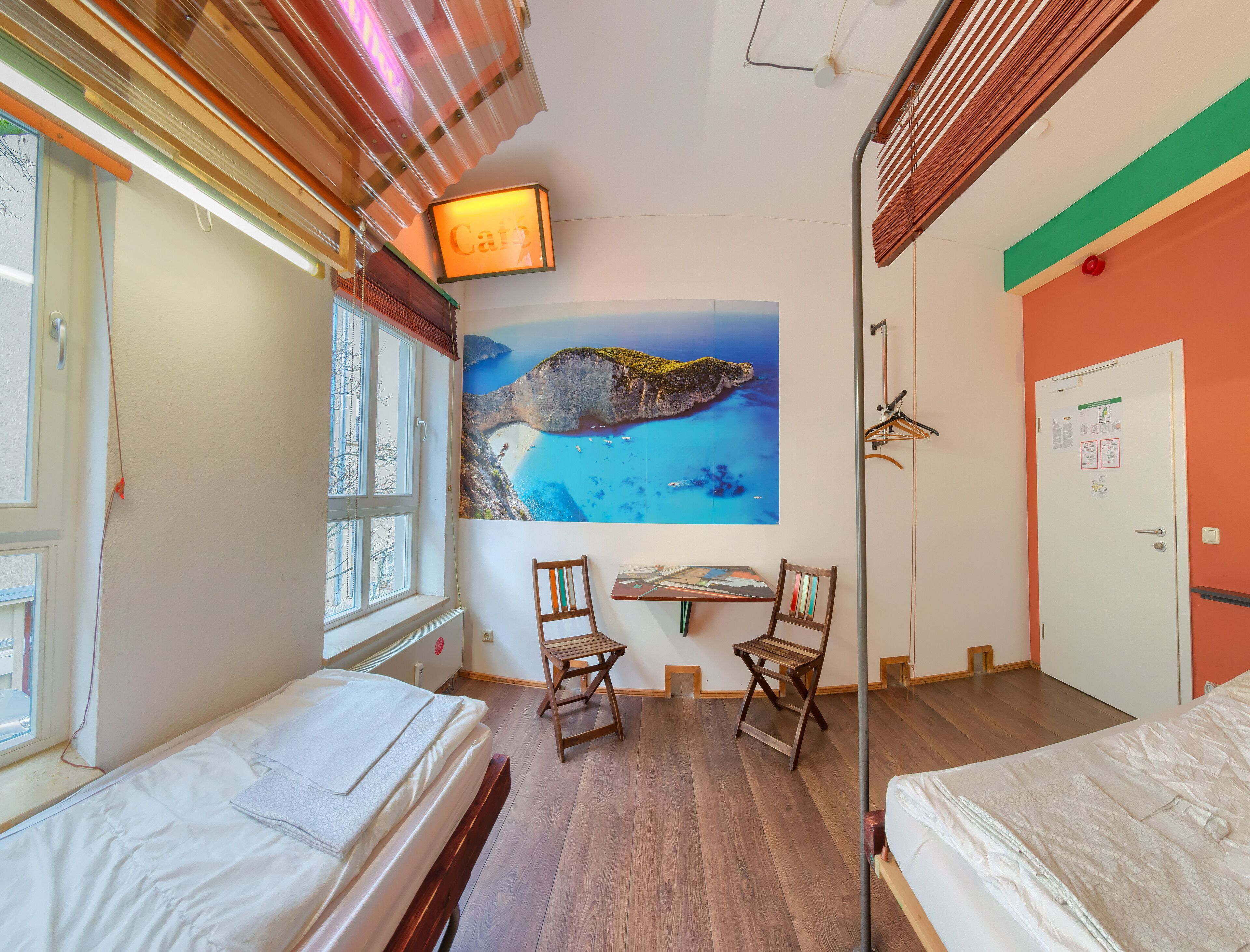 Habitación de hotel con dos camas, un escritorio y sillas, un cuadro de un paisaje costero y un cartel que dice 'Cafe'.