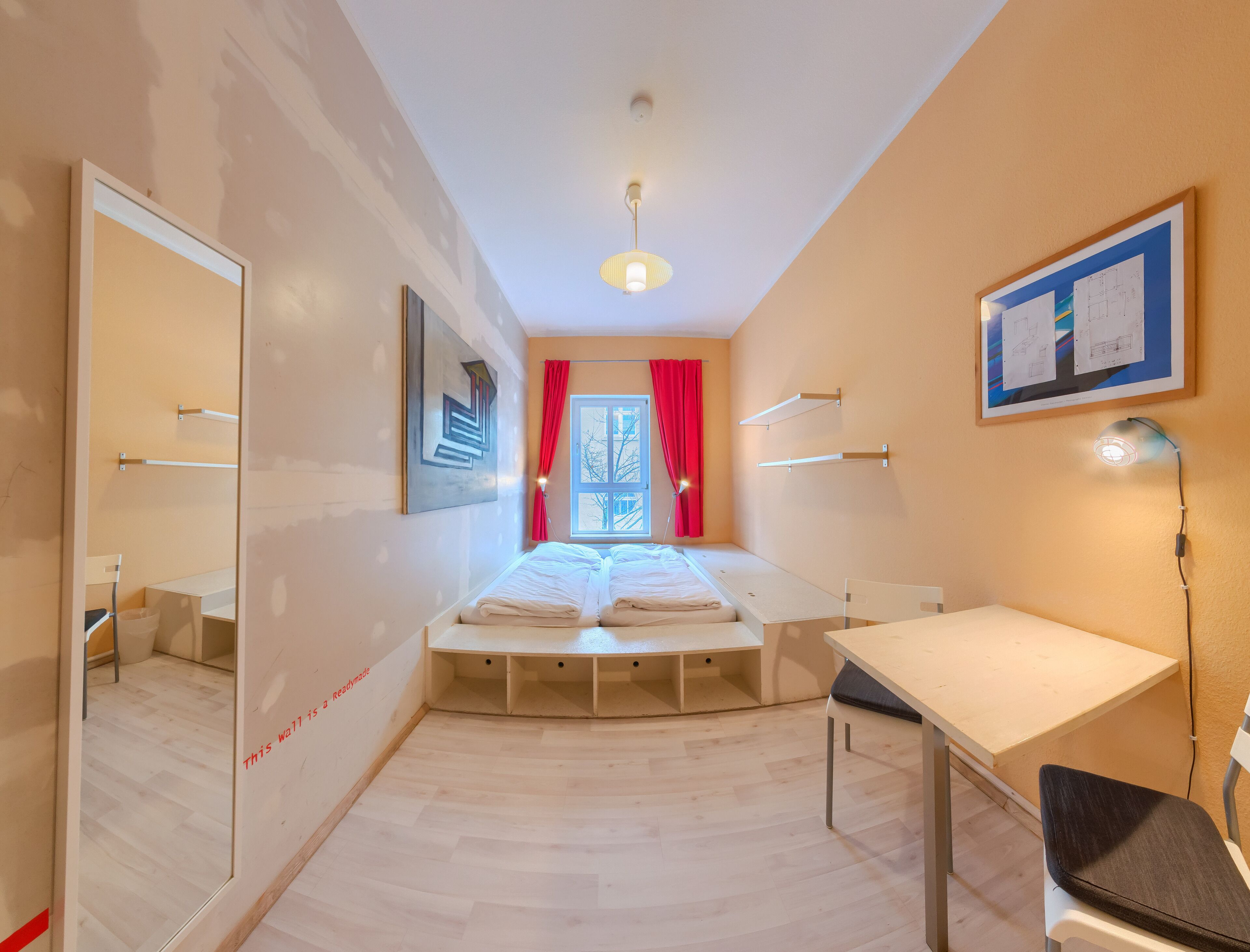 Habitación de hotel pequeña con cama, escritorio, silla, espejo y ventana con cortinas rojas.