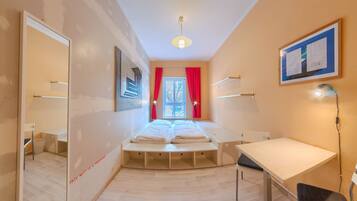 Habitación de hotel pequeña con cama, escritorio, silla, espejo y ventana con cortinas rojas.