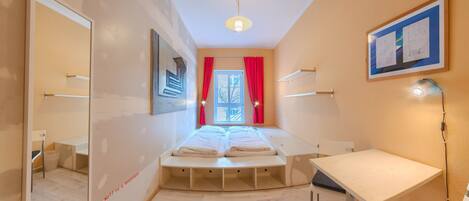 A compact hotel room with a bed, a desk, a chair, a mirror, and a window with red curtains.