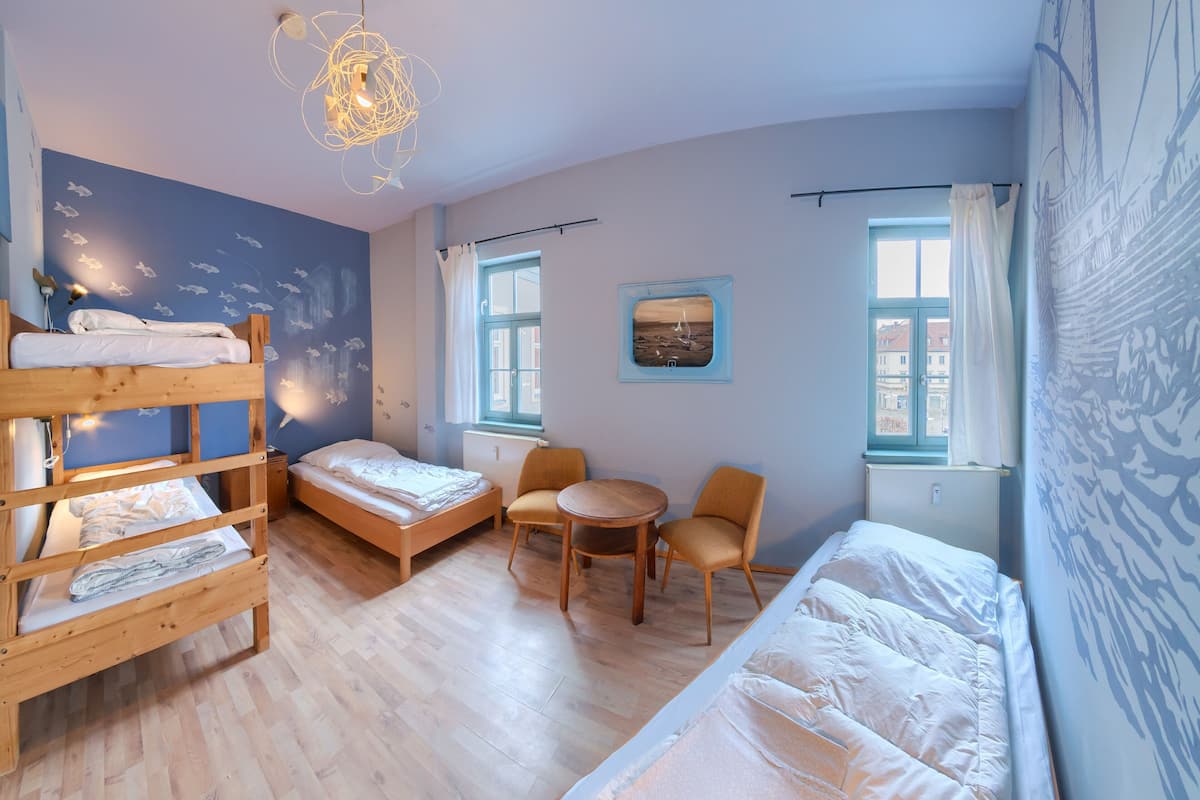 A room with bunk beds, a small table, chairs, and a view of buildings through a window.