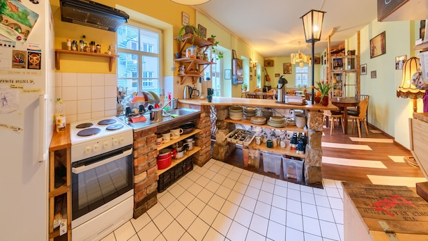 A kitchen with a stove, oven, and various kitchen items. There is a window, a hanging light fixture, and a shelf with kitchenware. - Labyrinth Hostel (Weimar)
