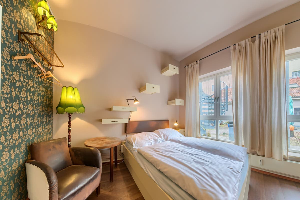 A hotel room with a bed, a chair, a small table, a lamp, and a window with curtains.