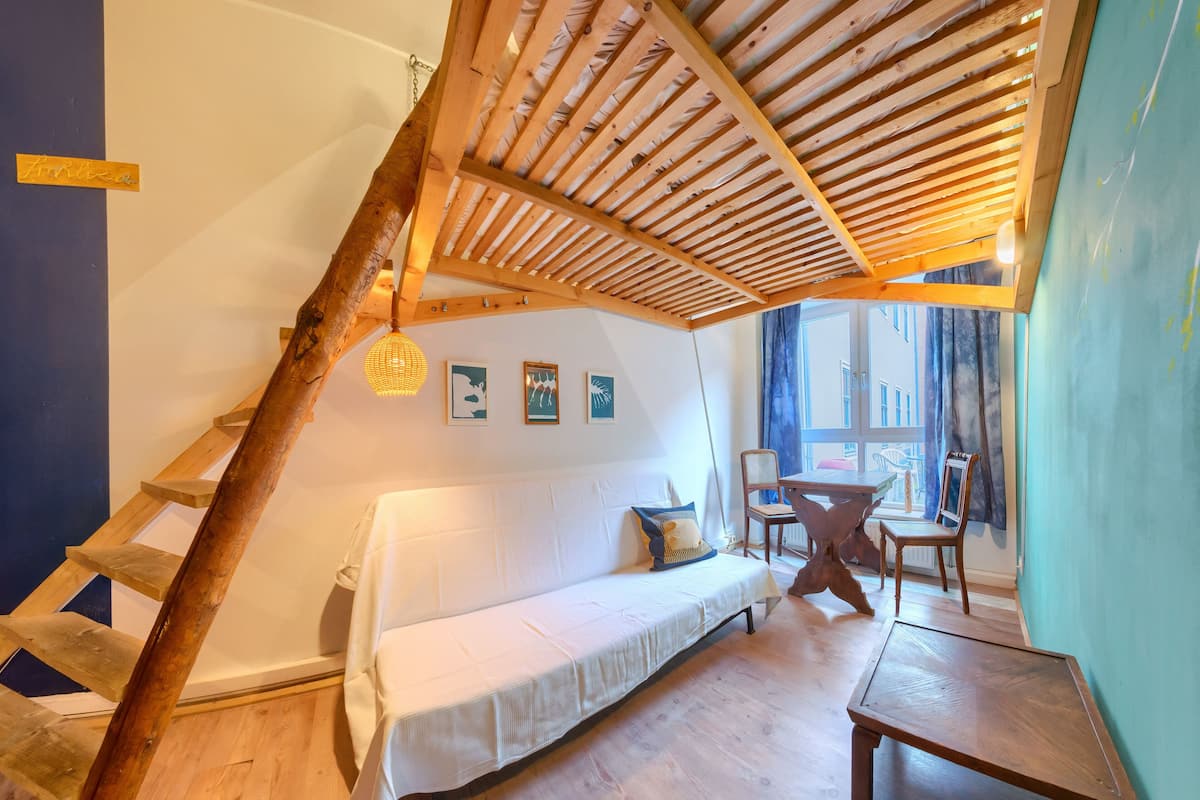 A cozy room with a wooden ladder, a white sofa, a wooden table, and a view of the street through large windows.