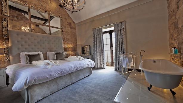 A spacious bedroom with a large bed, a classic clawfoot bathtub, and a stone accent wall. - Horsley Lodge Hotel (Derby)
