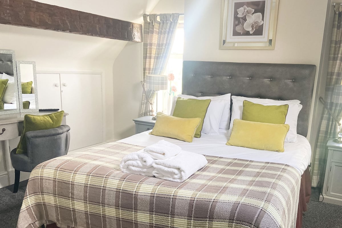 A neatly made bed with a plaid bedspread, two green and two yellow pillows, a white towel, a bedside table with a lamp, and a framed picture on the wall.