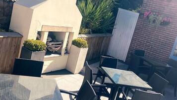 Un patio al aire libre con una parrilla empotrada, mobiliario con sillas y mesas, y un muro de ladrillo con plantas en macetas.
