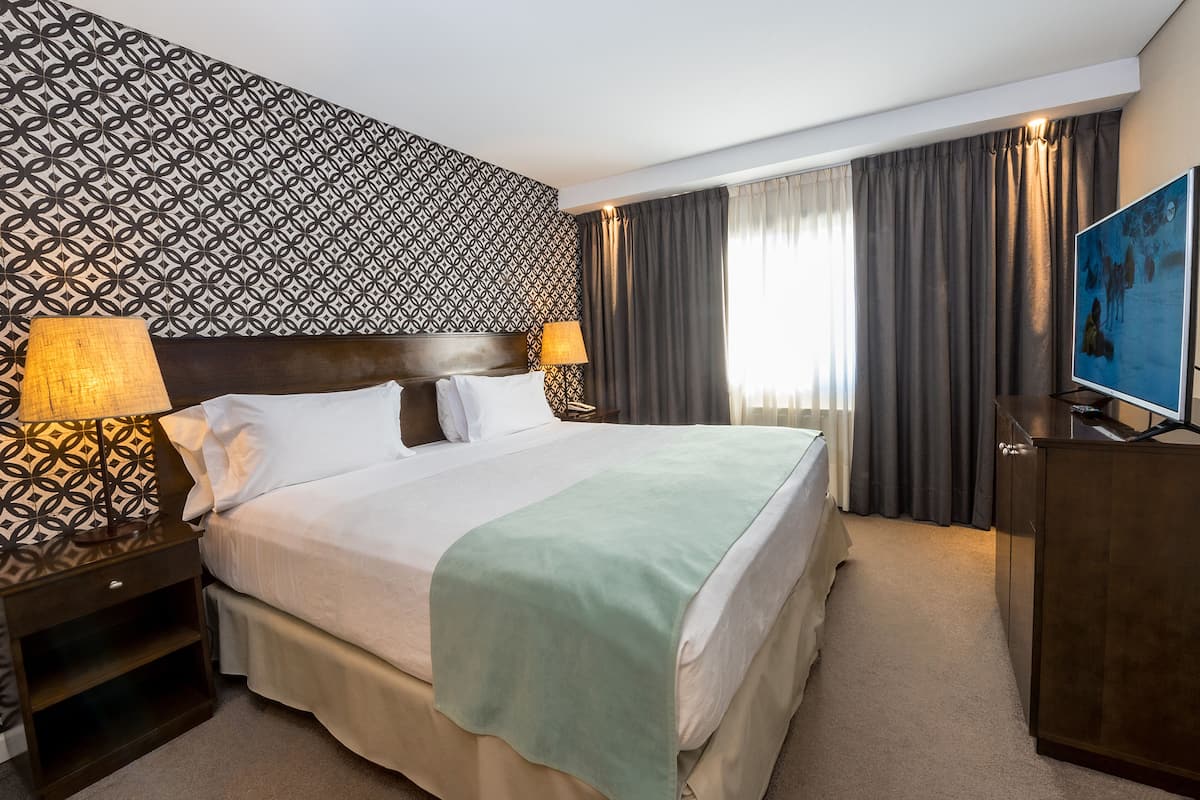 A hotel room with a large bed, two bedside tables with lamps, a TV, and a patterned wall.