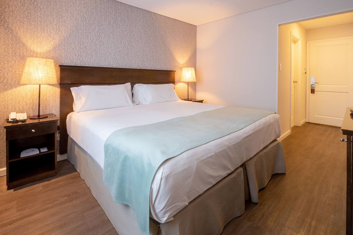 A neatly made bed with two pillows, a bedside table with a lamp, and a door to the right.
