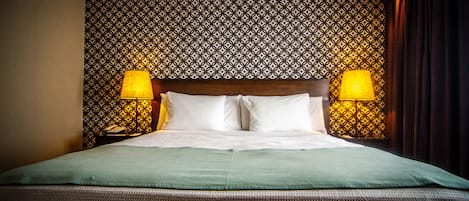 A hotel room with a patterned wallpaper, a wooden headboard, two bedside lamps, and a green bedspread.