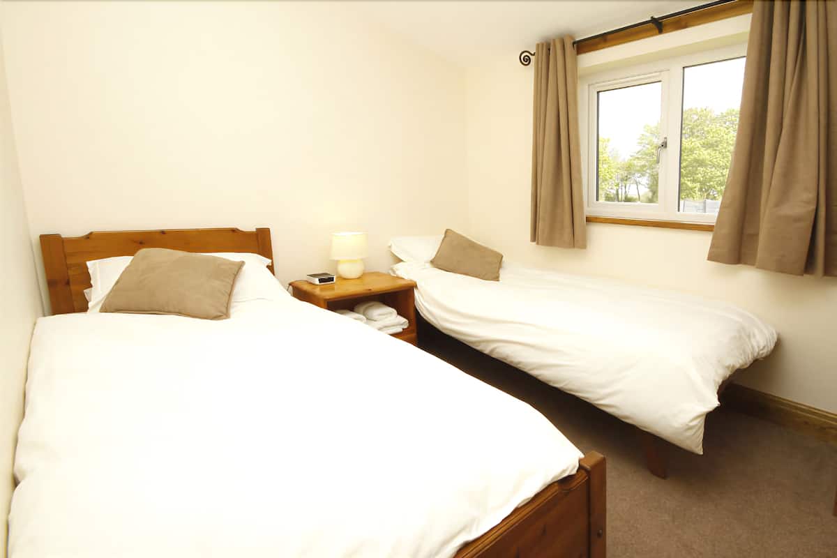 A bedroom with two single beds, a bedside table with a lamp, and a window with curtains.
