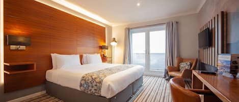 A hotel room with a large bed, a desk with a chair, a television, and a view of the sea.