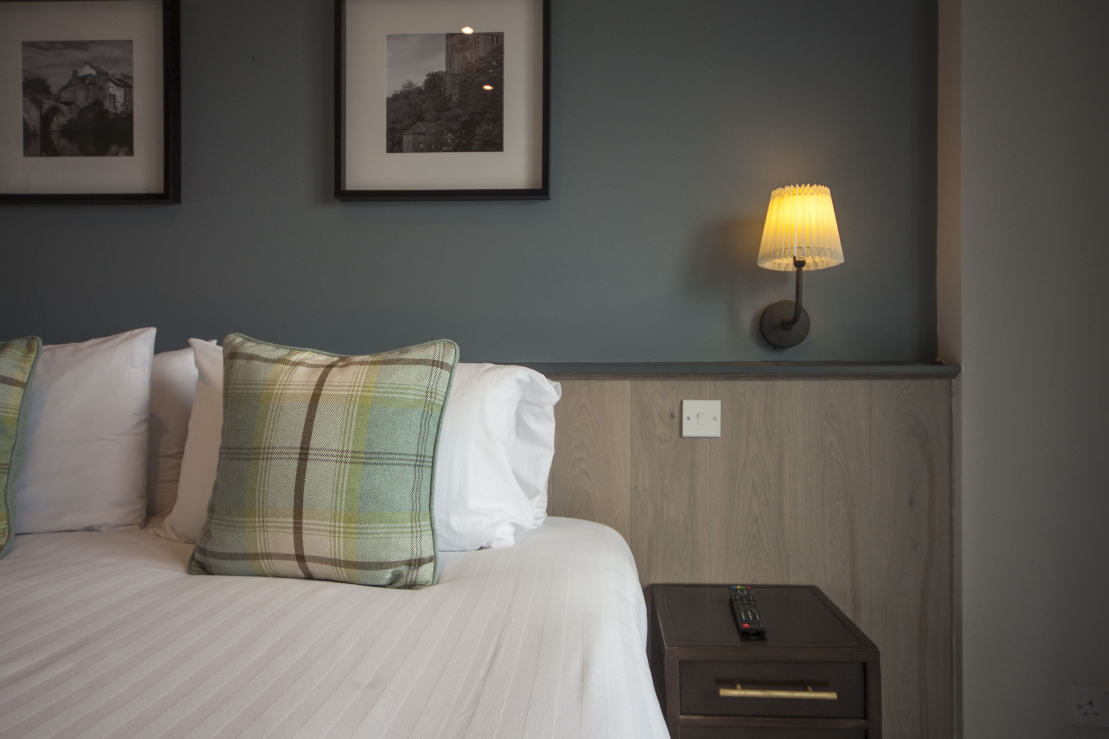 A neatly made bed with plaid and white pillows, a bedside lamp, and two framed pictures on the wall.