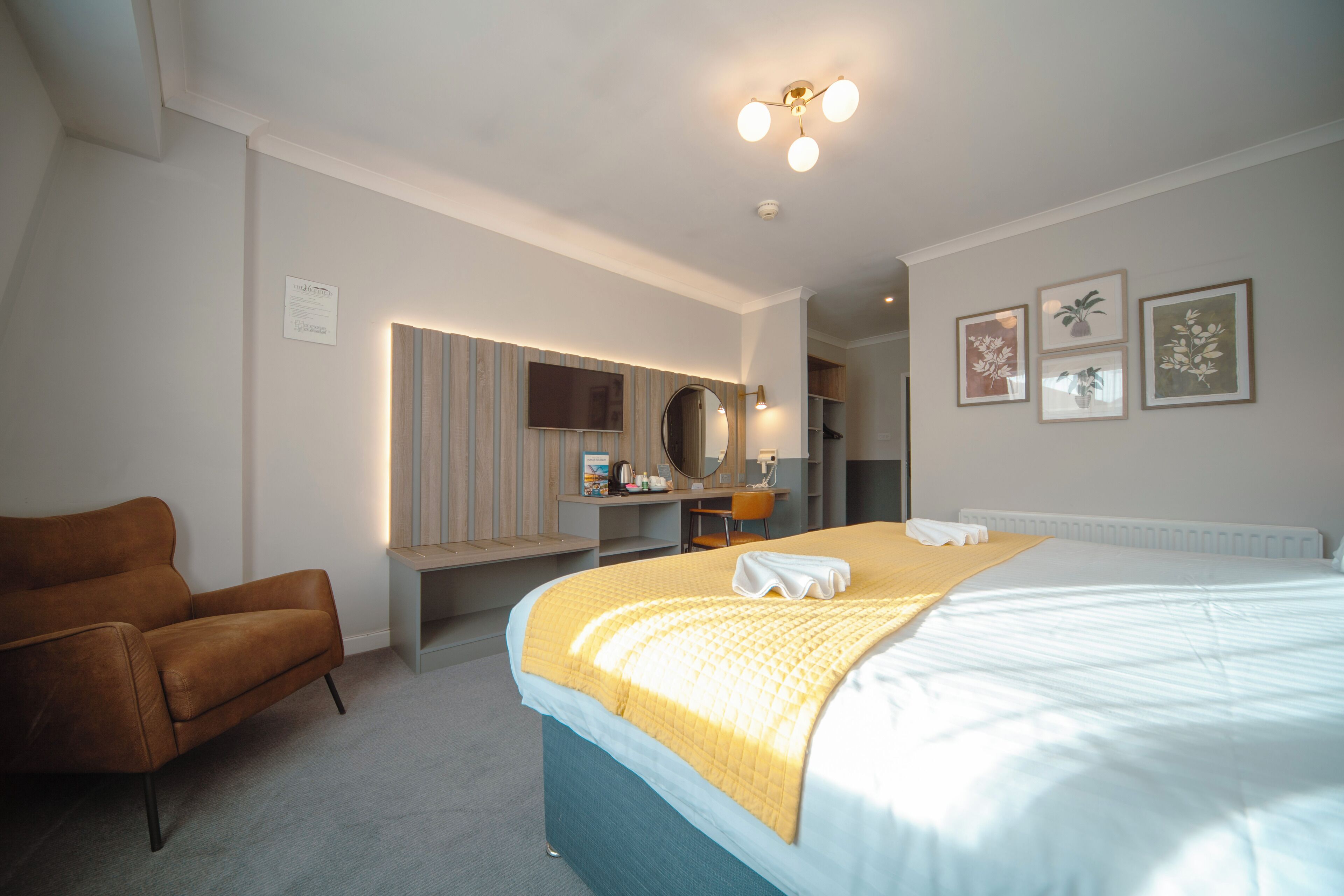 Une chambre d’hôtel avec un lit, une chaise, un bureau avec un ordinateur, un miroir et des tableaux encadrés au mur.