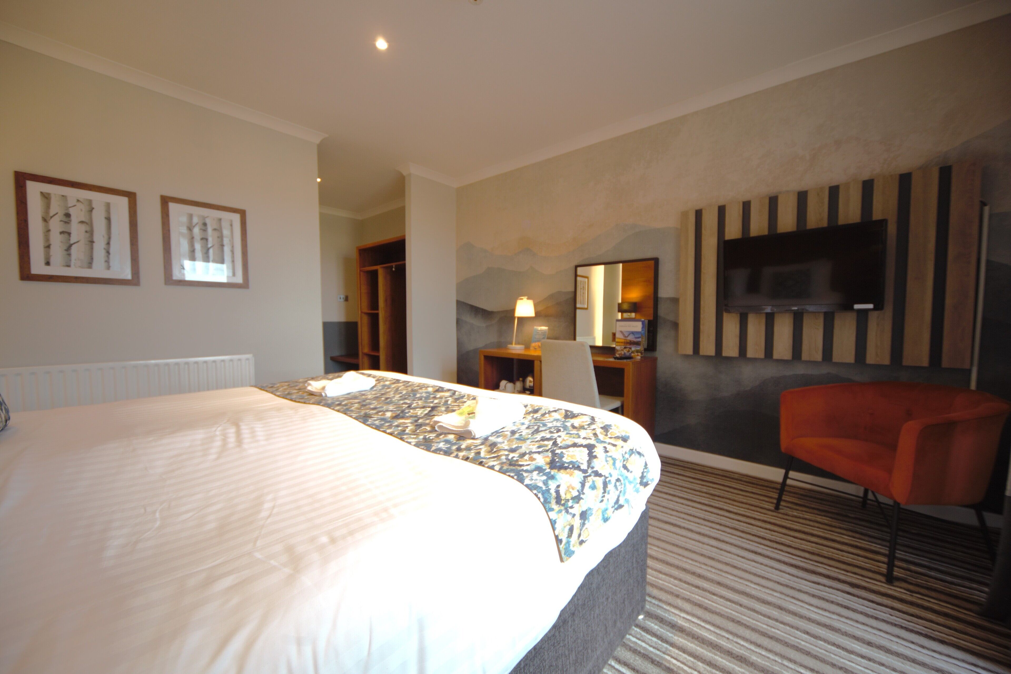 A hotel room with a large bed, a desk with a lamp, a TV mounted on the wall, and a red chair.