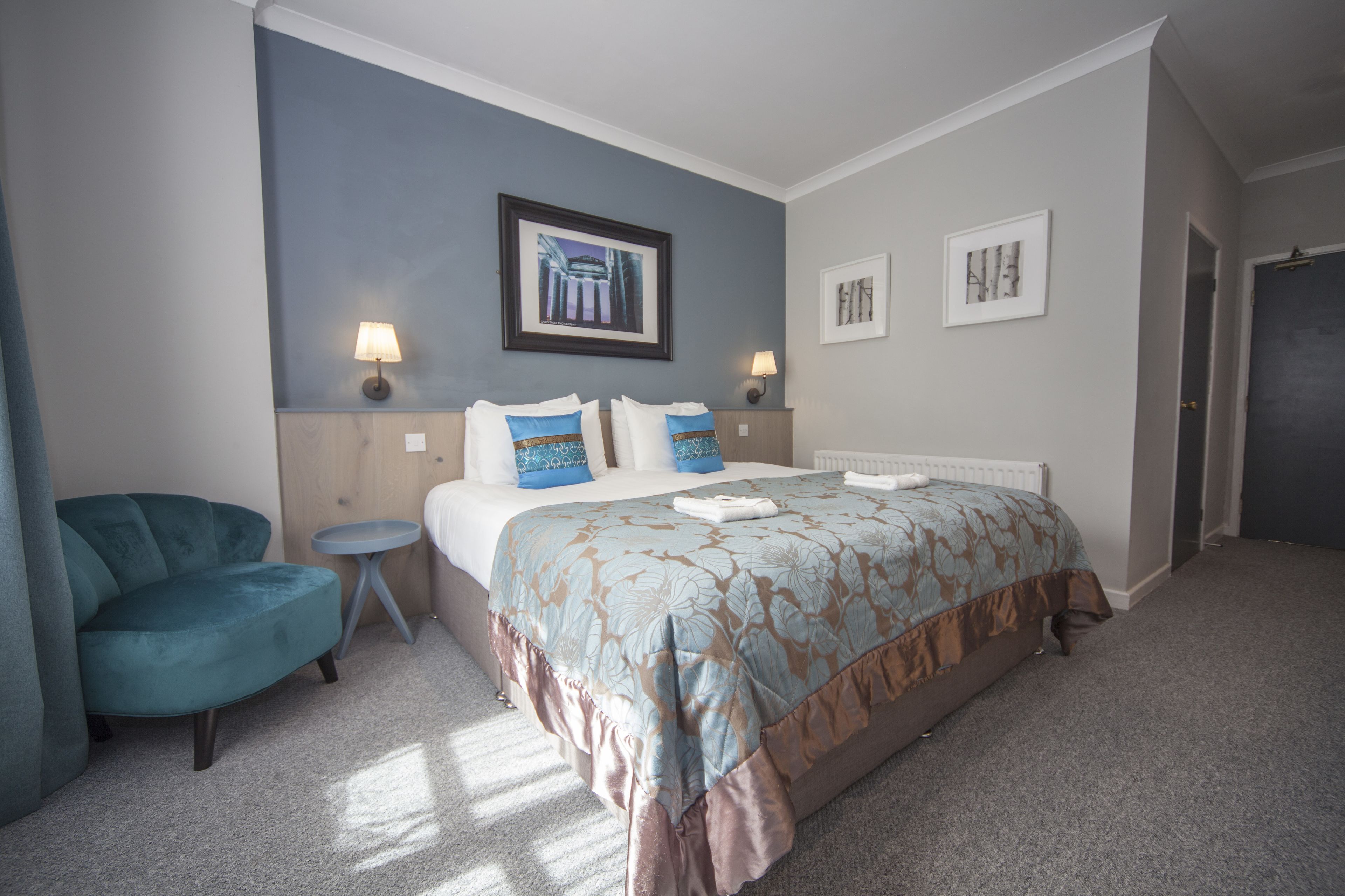 A hotel room with a bed, a small round table, a blue armchair, and two wall-mounted lamps.
